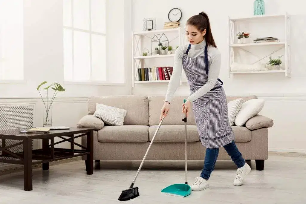 Efficient professional maid sweeping living room floor with broom and dustpan.