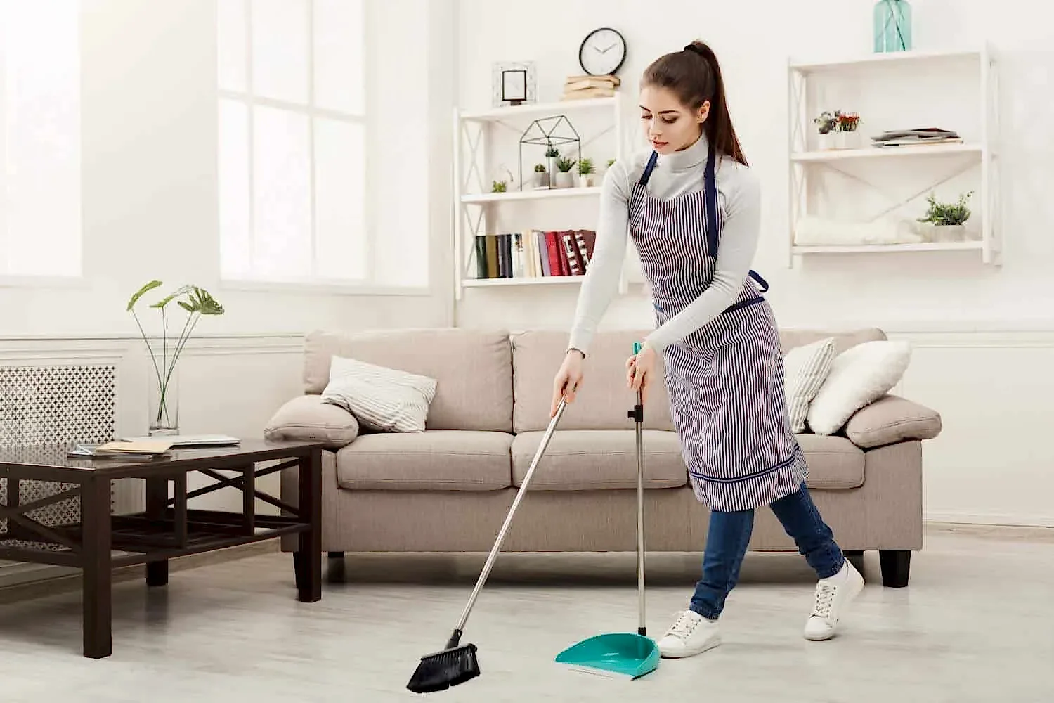 Efficient professional maid sweeping living room floor with broom and dustpan.